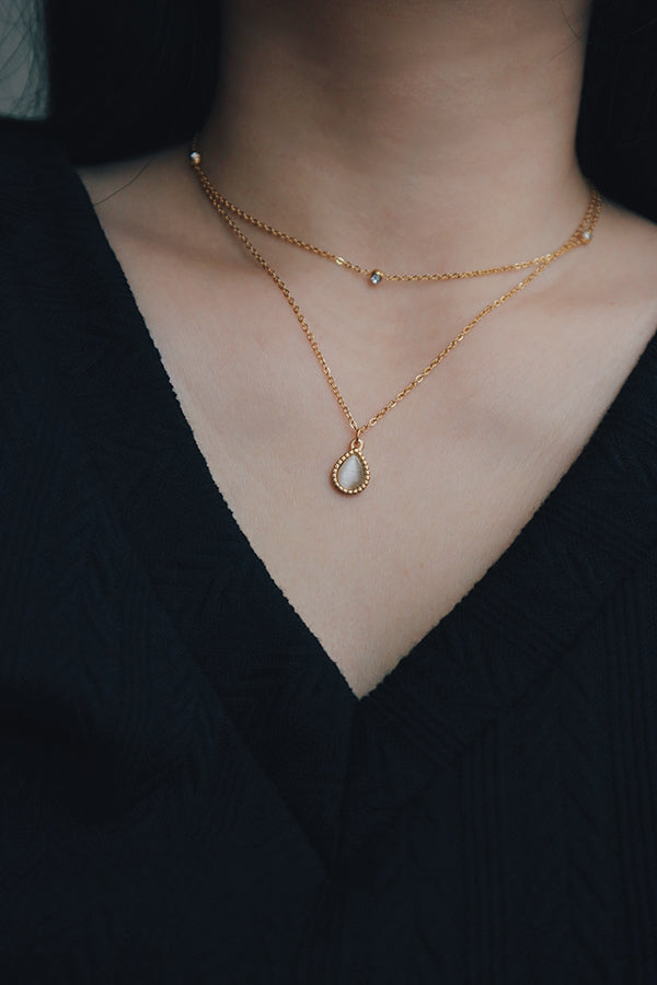 A layered necklace set featuring a dainty gold chain with a teardrop cat's eye pendant and a delicate chain with cubic zirconia stones, worn with a black V-neck top.