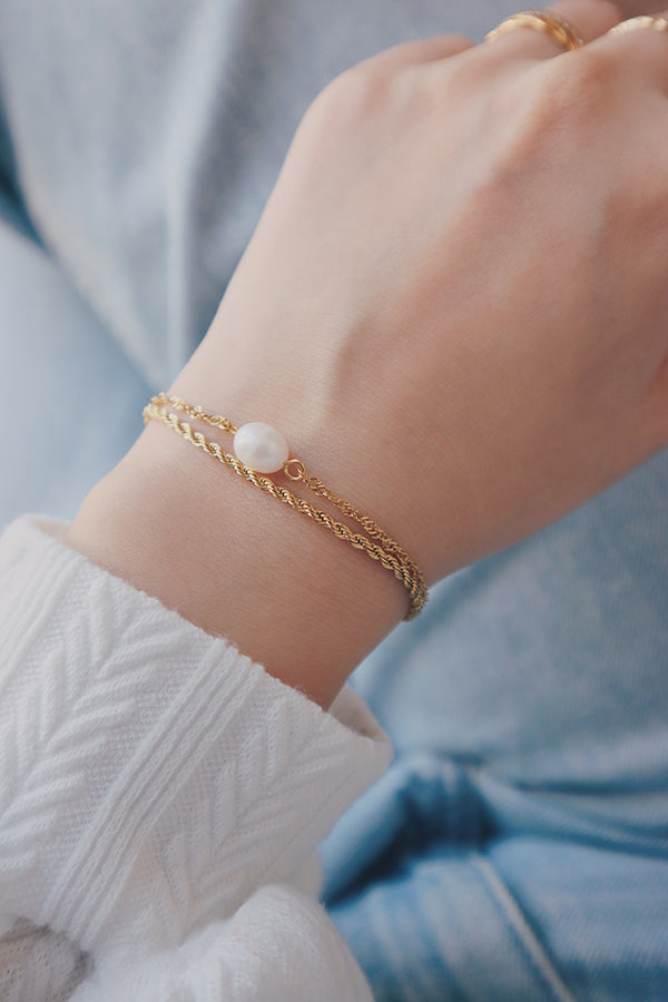 Close-up of a wrist adorned with a delicate layering bracelet featuring a single freshwater pearl and a twisted gold chain, styled with a white textured sleeve.