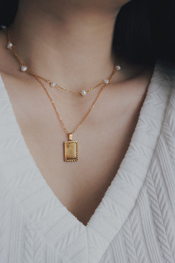 Close-up of a woman holding the Savita Sun pendant, which features a sunburst engraving, styled with a white textured top and layered gold necklaces.