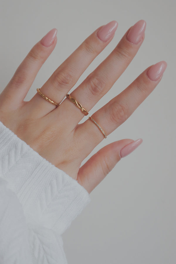 A close-up image of a womans hand wearing a delicate gold slim ring alongside other stackable rings. The ring features a subtle, wave-like design along its band, making it an elegant and minimalist piece perfect for mixing and matching with other jewelry.