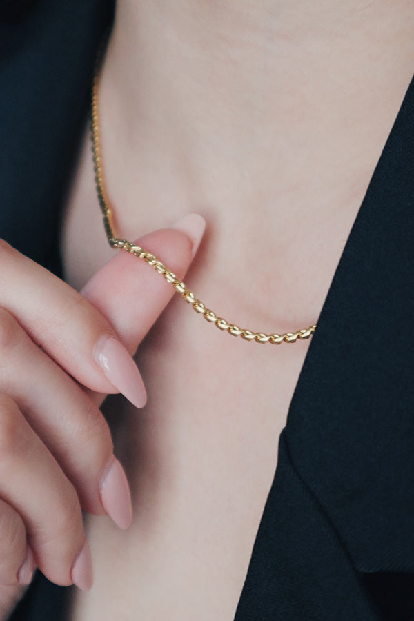 Close-up of a woman holding a 18K gold-plated rope chain necklace near her neck, paired with a black blazer, highlighting the luxurious texture and shine of the necklace.