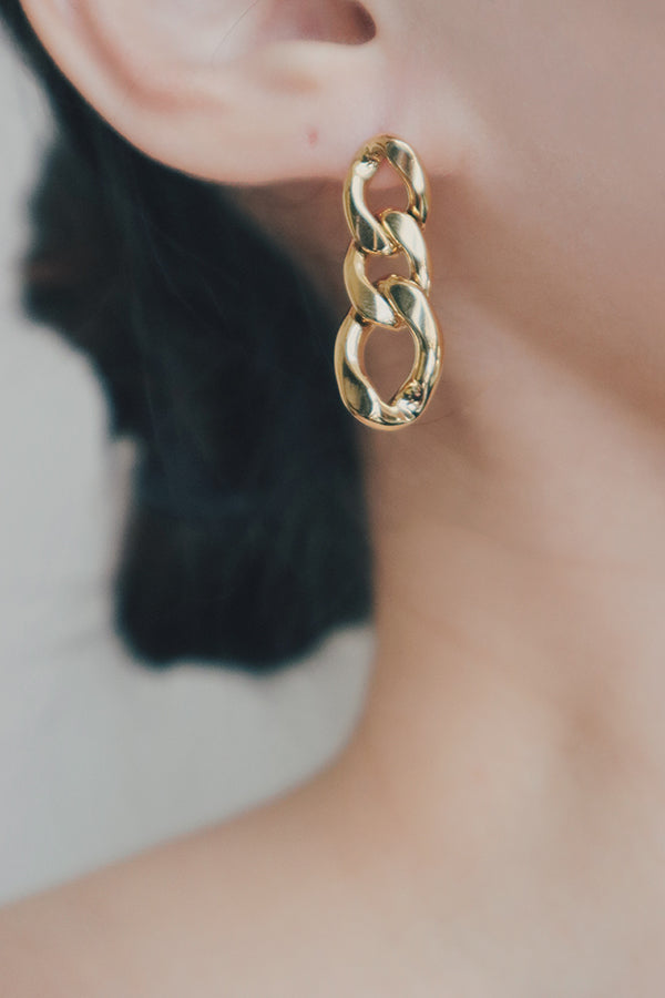 A close-up image of a womans ear wearing Cuban chain gold earrings. The earrings feature bold, interconnected links that form a chain, creating a statement piece that adds a touch of glamour and sophistication to her look.