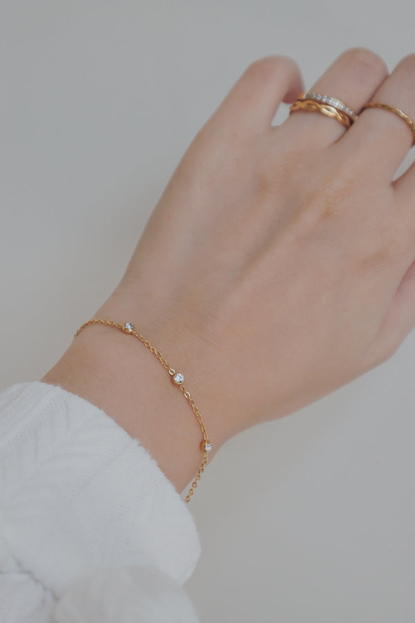 A minimalist gold bracelet featuring small, bezel-set cubic zirconia stones along a delicate chain, worn with a white textured sleeve.