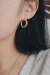 A detailed shot of a woman wearing a small, twisted texture gold hoop earring. The earring's surface features a delicate, ridged design that adds subtle texture and shine, making it a versatile accessory for everyday wear.