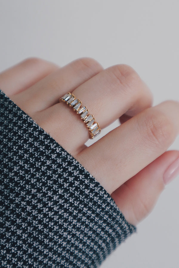 A close-up shot of a hand displaying a gold eternity ring with baguette-cut cubic zirconia stones. The ring's sleek and polished design features a continuous row of sparkling stones, set against a houndstooth-patterned sleeve that complements the ring's bold and elegant style.