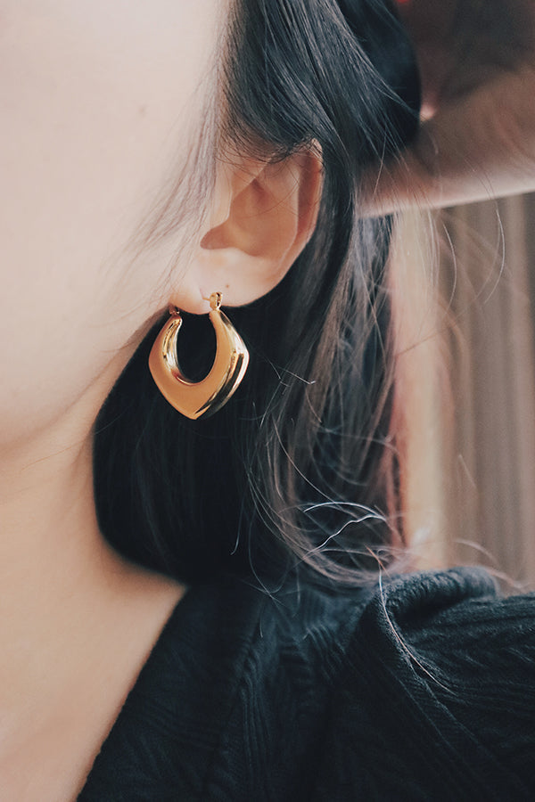 A close-up image of a woman modeling a large, square-shaped gold hoop earring. The earring features a sleek, polished finish, enhancing its bold and stylish design. The geometric shape adds a touch of modern elegance to the look.