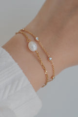 Close-up of a wrist wearing the SH & Co. Maeve pearl bracelet featuring a freshwater pearl and a delicate gold chain, styled with a white top and gold rings for a sophisticated finish.