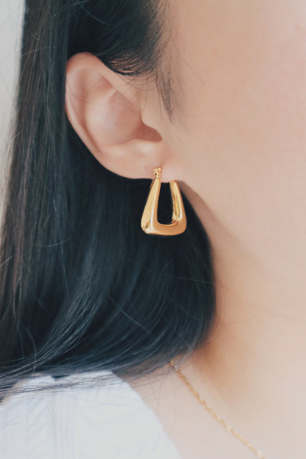 A detailed image of a woman wearing gold trapezoid hoop earrings. The earrings are trapezoid-shaped with a smooth, shiny finish that reflects light beautifully, making them a sophisticated addition to her jewelry collection.