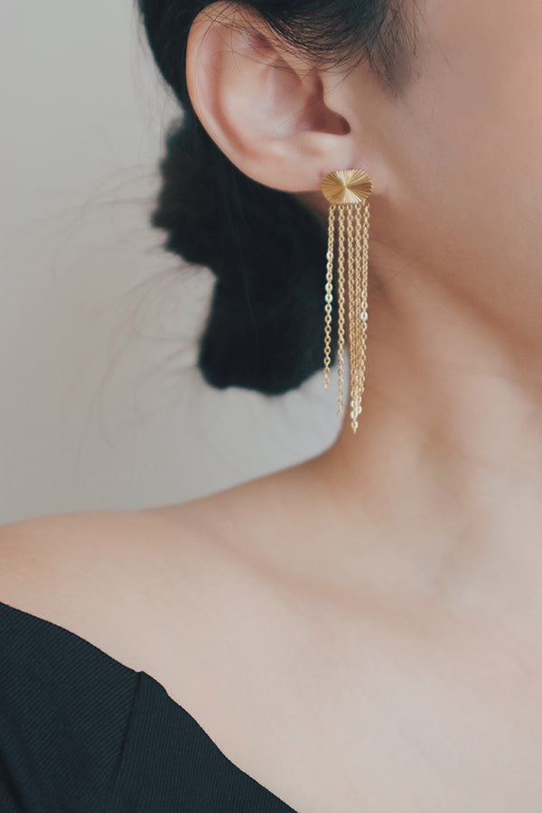 A close-up image of a woman's ear adorned with gold tassel earrings. The earrings have a sunburst design at the top and several gold chains that fall delicately, offering a stylish and contemporary look.