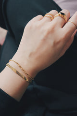 Close-up of a womans wrist wearing the SH & Co. Sara rope bracelet in 18K gold-plating, styled with a black outfit and gold rings for a chic, coordinated look.