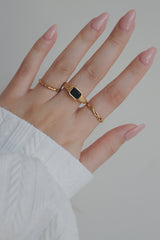 A detailed image of a gold ring featuring a black cubic zirconia stone set in a rectangular frame. The ring's modern and chic design makes it a versatile accessory for stacking or wearing alone for a minimalistic statement.