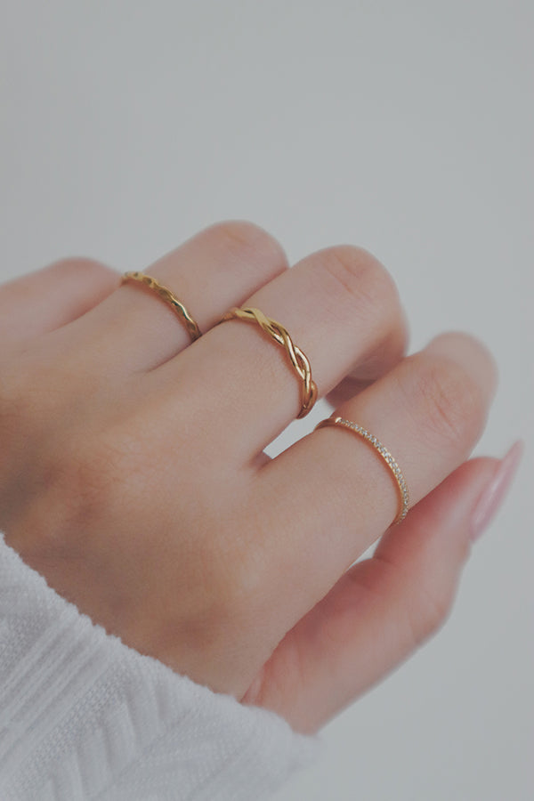 A detailed shot of a gold woven ring on a woman's hand. The ring is part of a stylish stack, with its braided design adding texture and depth to the overall look. The polished gold finish enhances its versatility as a layering piece.