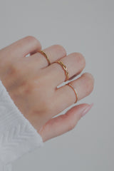 A close-up image of a womans hand wearing a gold woven ring alongside other stackable rings. The ring features an intricate braided design, with smooth, interwoven strands creating a timeless and elegant look. The hypoallergenic material makes it suitable for sensitive skin.