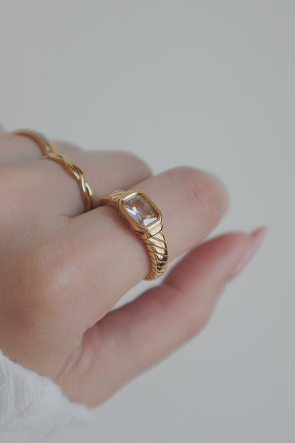 A detailed shot of a womans hand wearing a gold ring with a clear cubic zirconia stone. The rings rectangular stone and intricate band design make it an elegant choice for stacking with other rings or as a statement piece on its own.