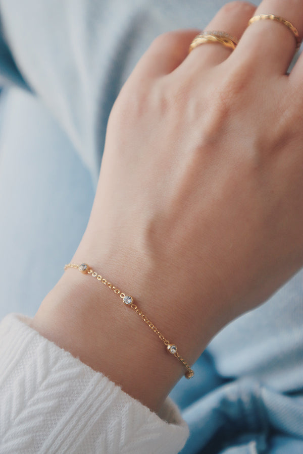 A dainty stainless steel bracelet featuring evenly spaced cubic zirconia stones, worn on the wrist along with gold rings, styled with a casual white and blue outfit.