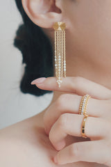 A close-up photo of a woman wearing gold tassel earrings. The earrings feature a sunburst pattern on the stud, with multiple fine chains that hang down, adding movement and sophistication to her look.