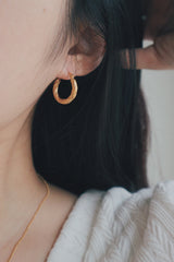 A close-up image of a woman's ear wearing a small gold hoop earring with a twisted texture design. The earring has a rich, golden hue that contrasts with the model's dark hair, creating an elegant and timeless look.