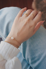 A wrist wearing a tarnish-free gold bracelet with a single freshwater pearl, paired with a white textured top and light blue jeans, highlighting a stylish and casual look.