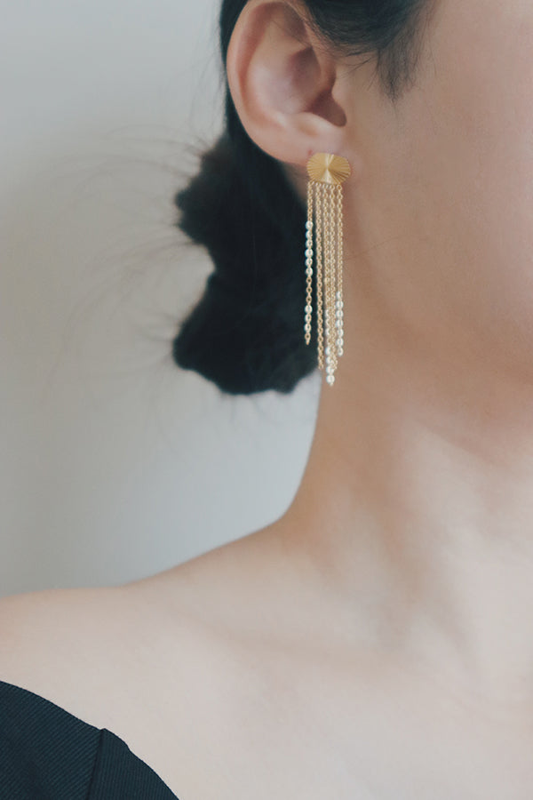 A close-up shot of a woman wearing gold tassel earrings. The earrings have a sunburst design on the stud, with several strands of fine gold chains that dangle elegantly, making them a perfect statement piece for any occasion.